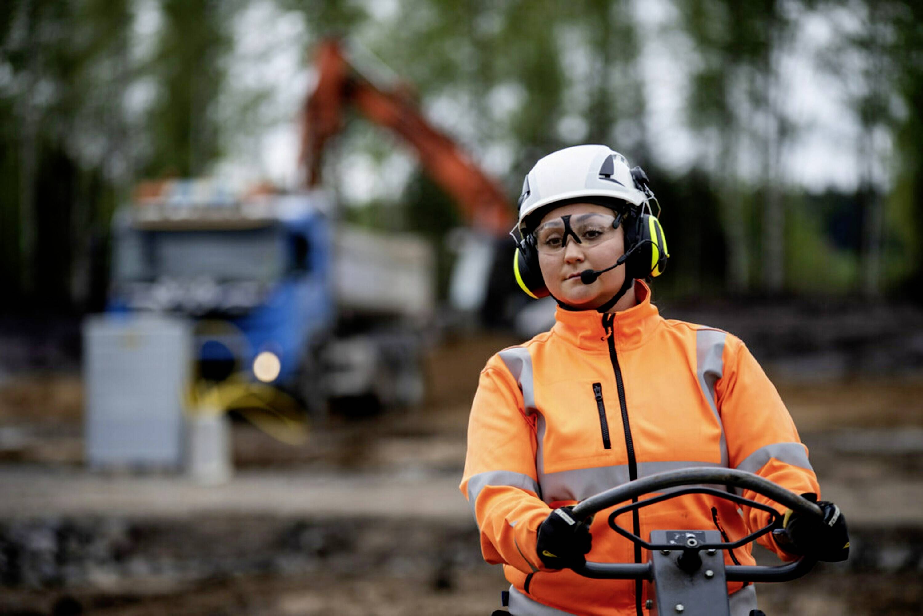 A person in orange protective clothing and a helmet is operating heavy machinery on a construction site; a lorry is blurred in the background.