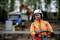 A person in orange protective clothing and a helmet is operating heavy machinery on a construction site; a lorry is blurred in the background.