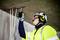 A worker wearing a hard hat and high-visibility vest is checking cables emerging from the ceiling. He is wearing gloves and ear protection.