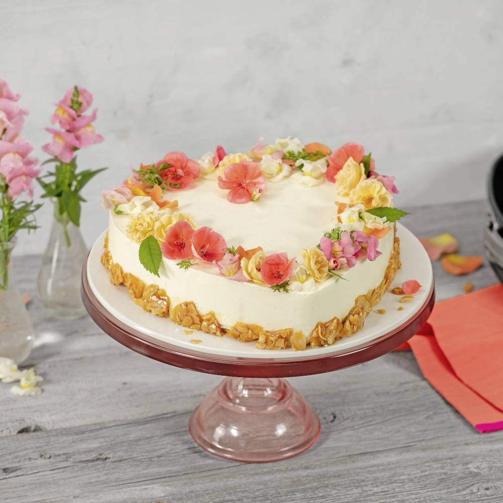 A round cake with white icing decorated with colourful flowers sits on a glass cake stand. Flowering blooms are placed in vases nearby.