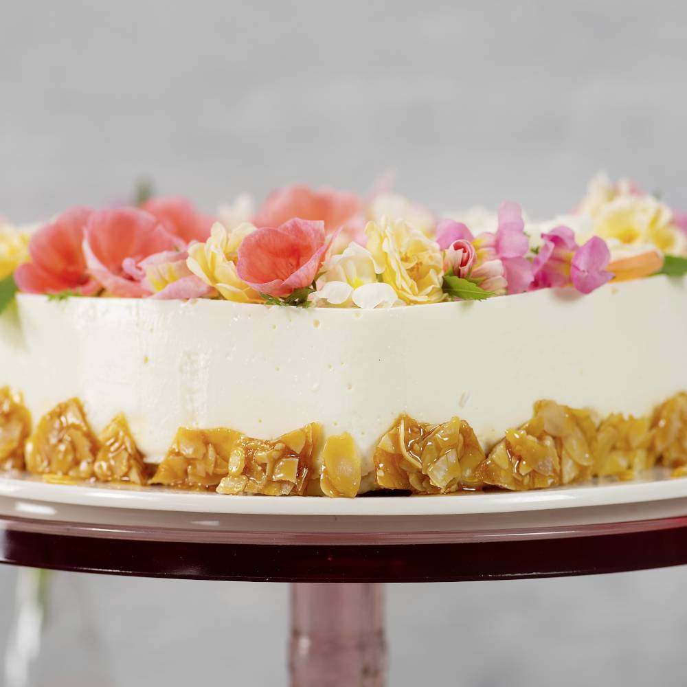 A round cake with cream-white icing, decorated with red and yellow flowers on top and candied nuts around the sides.