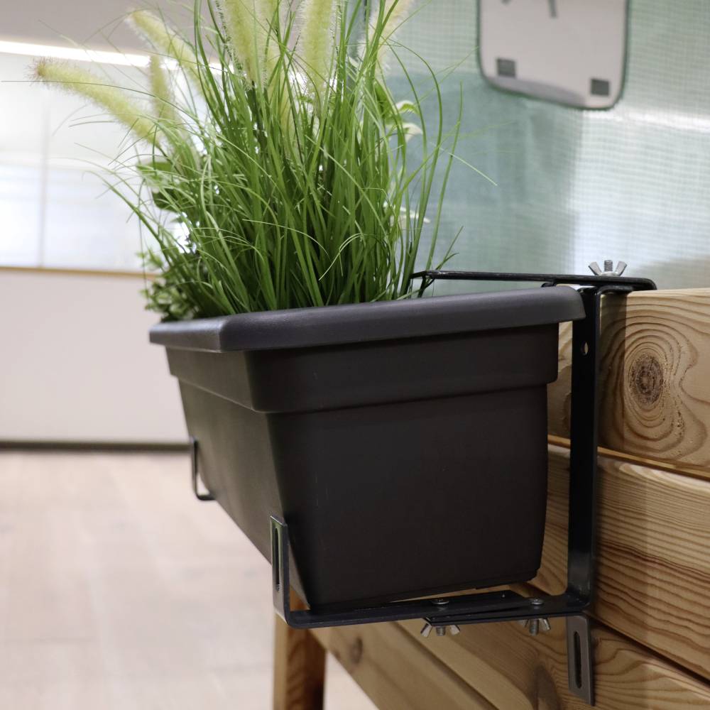 A black planter with green grass is attached to a wooden balcony railing. Tiles and a window are visible in the background.
