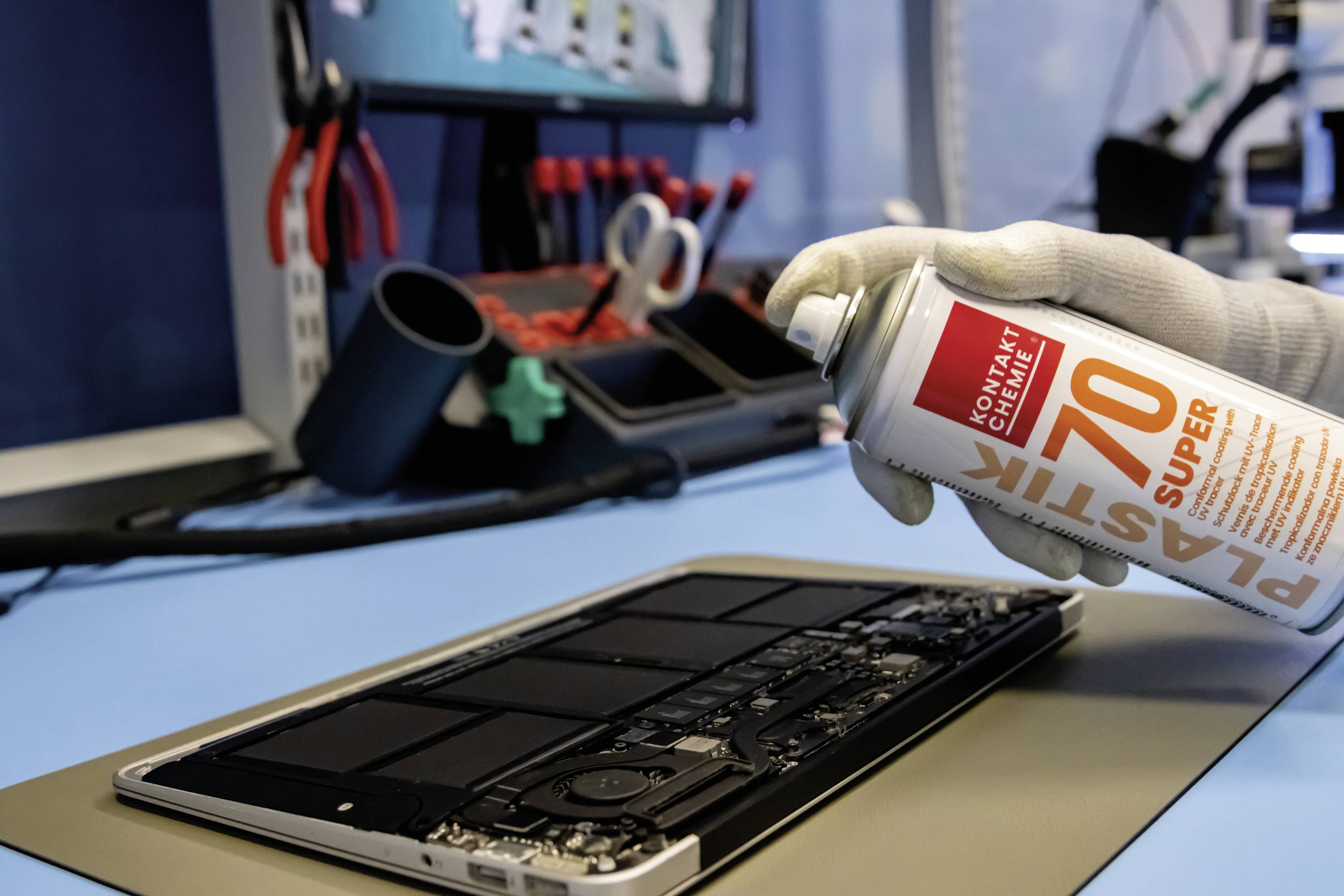 A hand in a glove sprays cleaner onto an open laptop on a table with tools. Repair scene in a workshop.