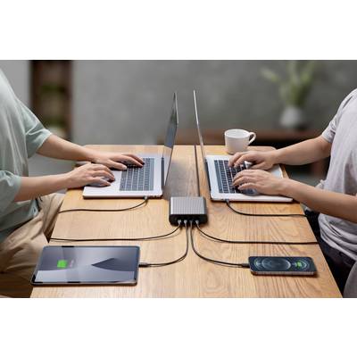 Two people are working on laptops at a table, connected to a charger. A tablet and a smartphone are being charged simultaneously.