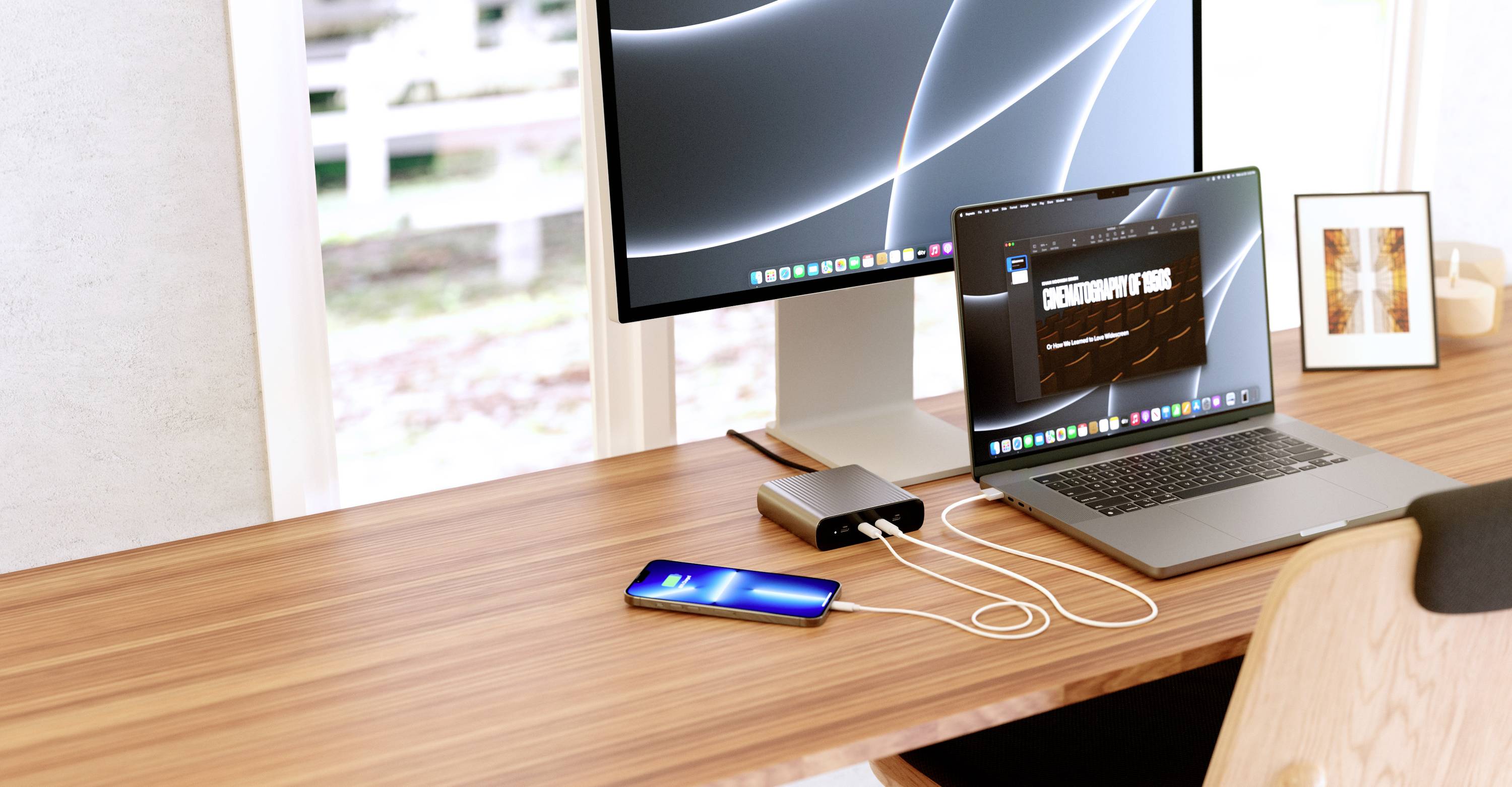 Workplace with a laptop and smartphone, both connected to a charging device. A monitor displays abstract lines on a black background.