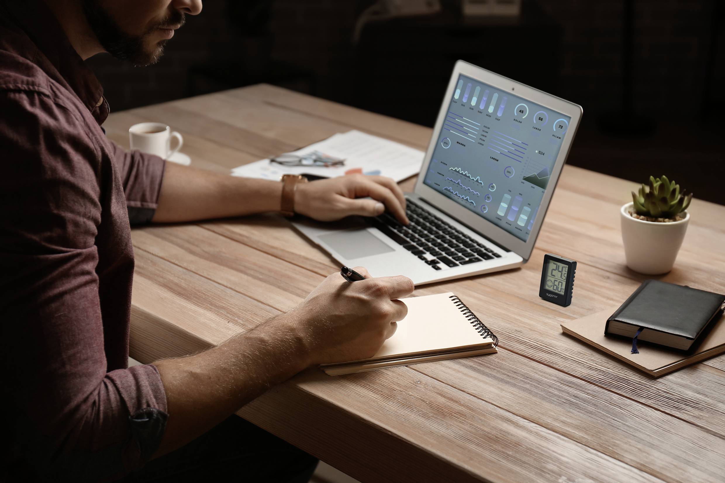 A man is sitting at a wooden table, using a laptop with data on the screen and making notes in a notebook.
