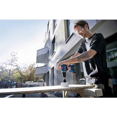 A man is drilling a hole in a wooden board on a construction site using an electric drill, wearing safety glasses.