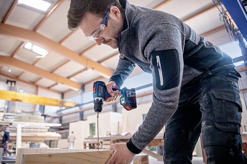 A man wearing safety glasses is drilling a hole in a piece of wood using an electric screwdriver in a woodworking workshop. Wooden beams can be seen in the background.