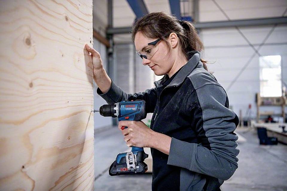 A female worker in a workshop is drilling a hole in a wooden board using a blue cordless drill. She is wearing safety glasses.
