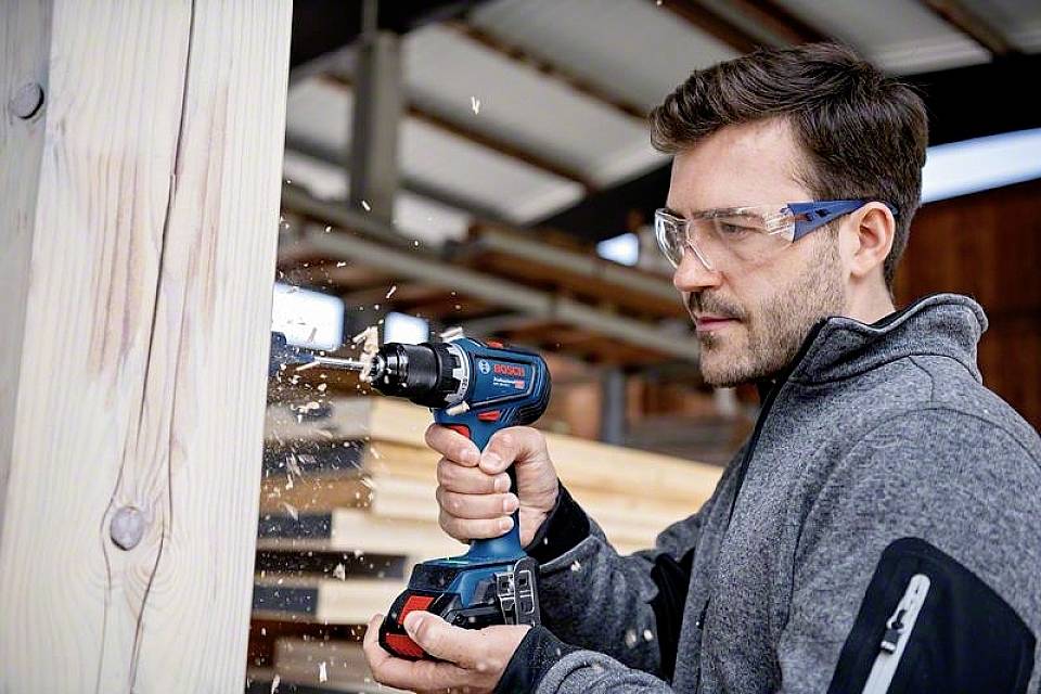 A man is wearing safety glasses and drilling a hole into a wooden beam with a cordless drill. Stacks of wood can be seen in the background.