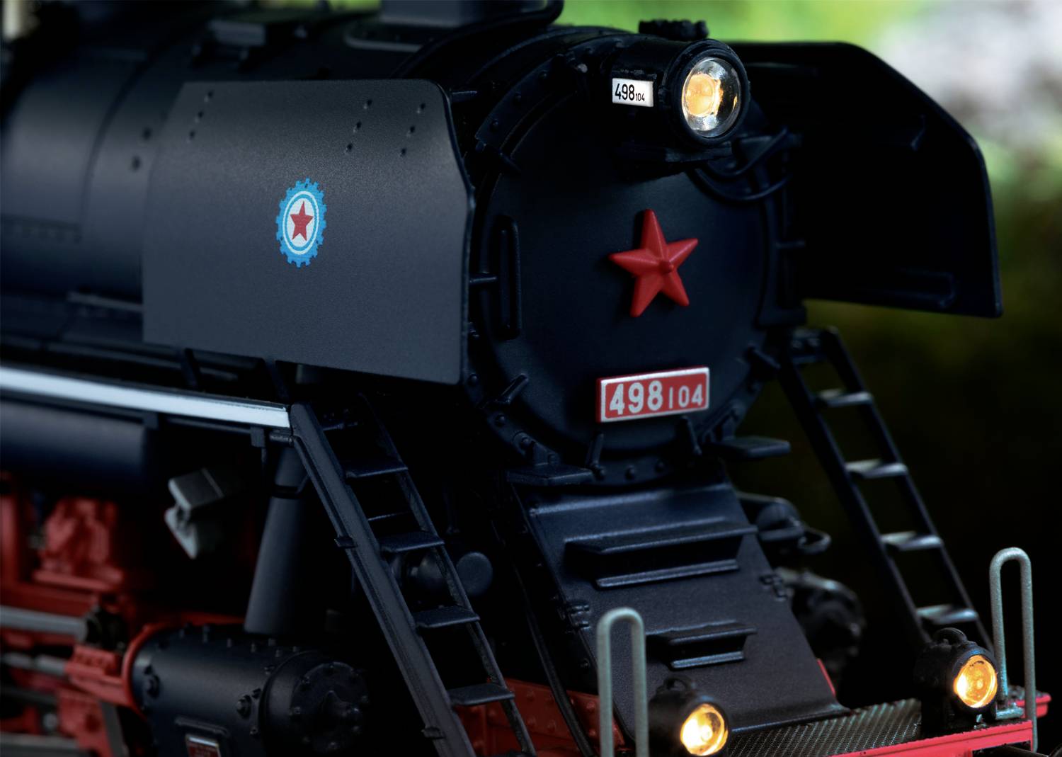 Black steam locomotive with red star emblem and the number '498.104' on the front. Illuminated headlamps, surrounded by trees.