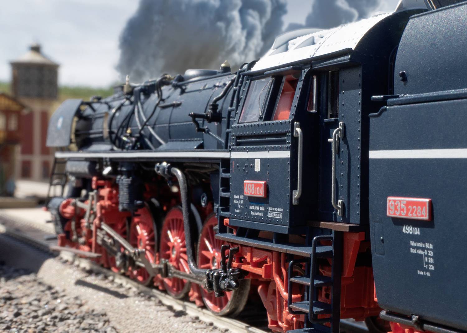 A steam locomotive travels along tracks, with white smoke billowing from its chimney. A building can be seen in the background.
