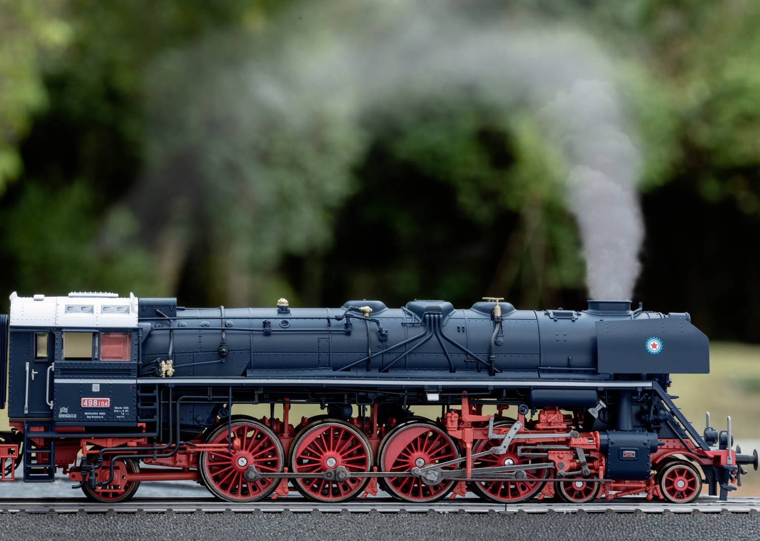 A black steam locomotive with red wheels travels along a track against a blurred, green background. Smoke rises from its chimney.