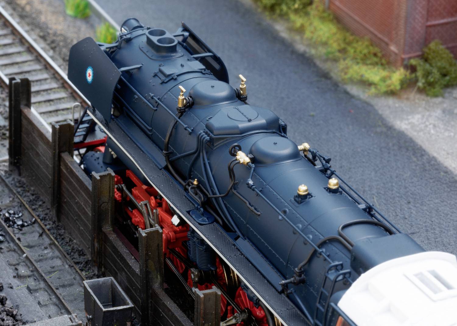 A black model steam locomotive stands on the tracks in a miniature landscape with plants and sheds in the background.