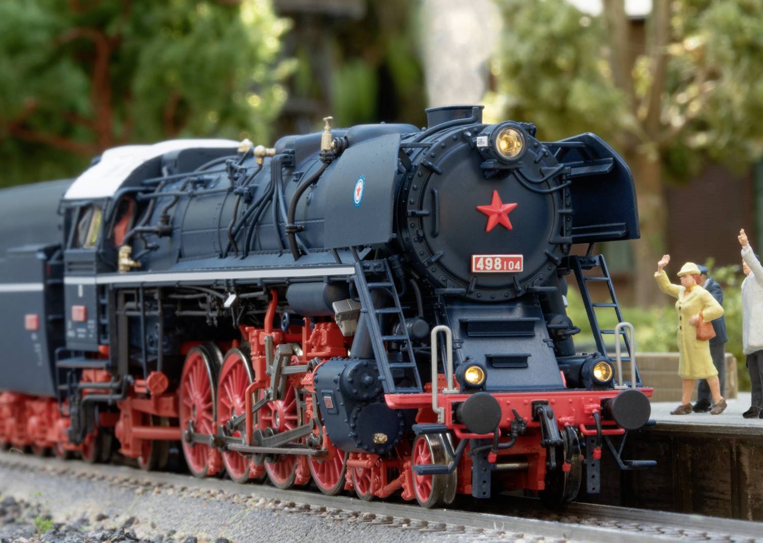 A black steam locomotive with red wheels travels along a railway track. Figures stand beside the locomotive, waving cheerfully.