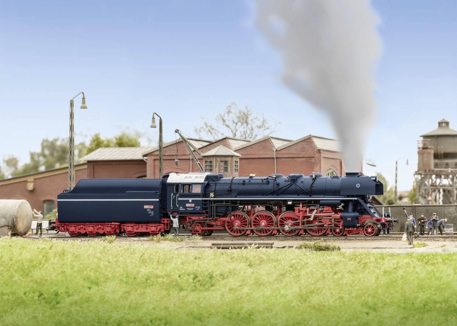 A blue model train with a red chassis sits on a track in front of an old, brick-red building. Smoke rises from the chimney.