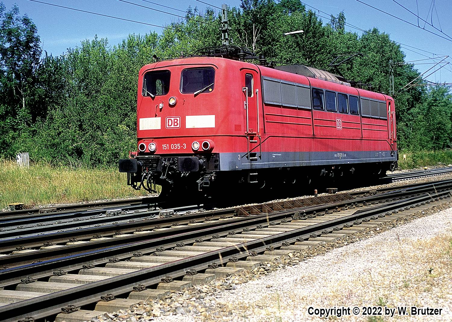 Red train of Deutsche Bahn on a railway track in a rural setting on a sunny day. Trees in the background.