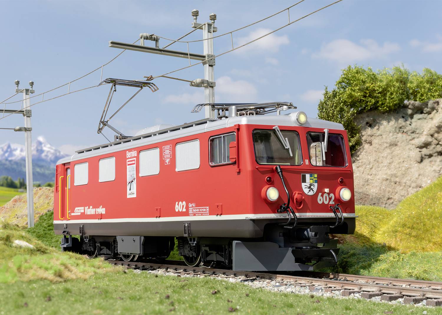 Red train travels on a narrow-gauge railway through a verdant mountain landscape. Overhead lines and electricity pylons line the route.