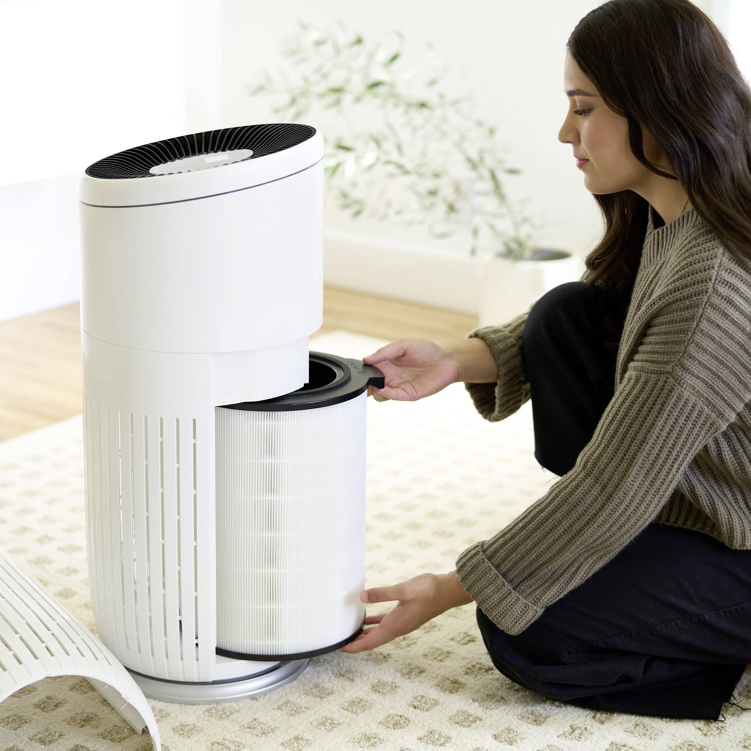 A woman is sitting on the floor and cleaning or replacing the filter of a white air purifier in a bright room.