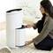 A woman is sitting on the floor and cleaning or replacing the filter of a white air purifier in a bright room.