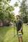 A man is watering a tree in the garden with a handheld device. Houses and trees are visible in the background.