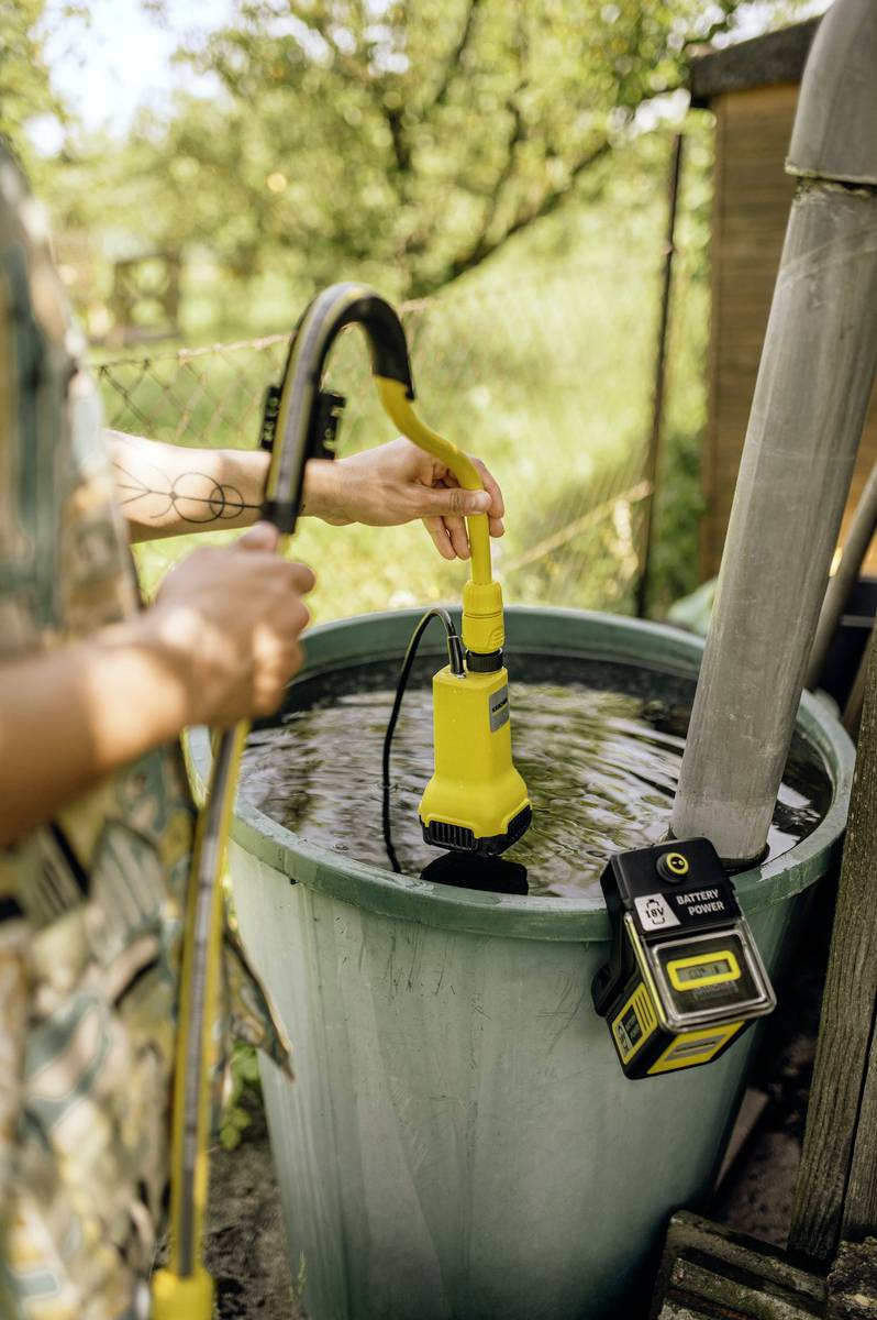 A person is using a yellow water pump to drain a full green water tank outdoors. It is a sunny day.