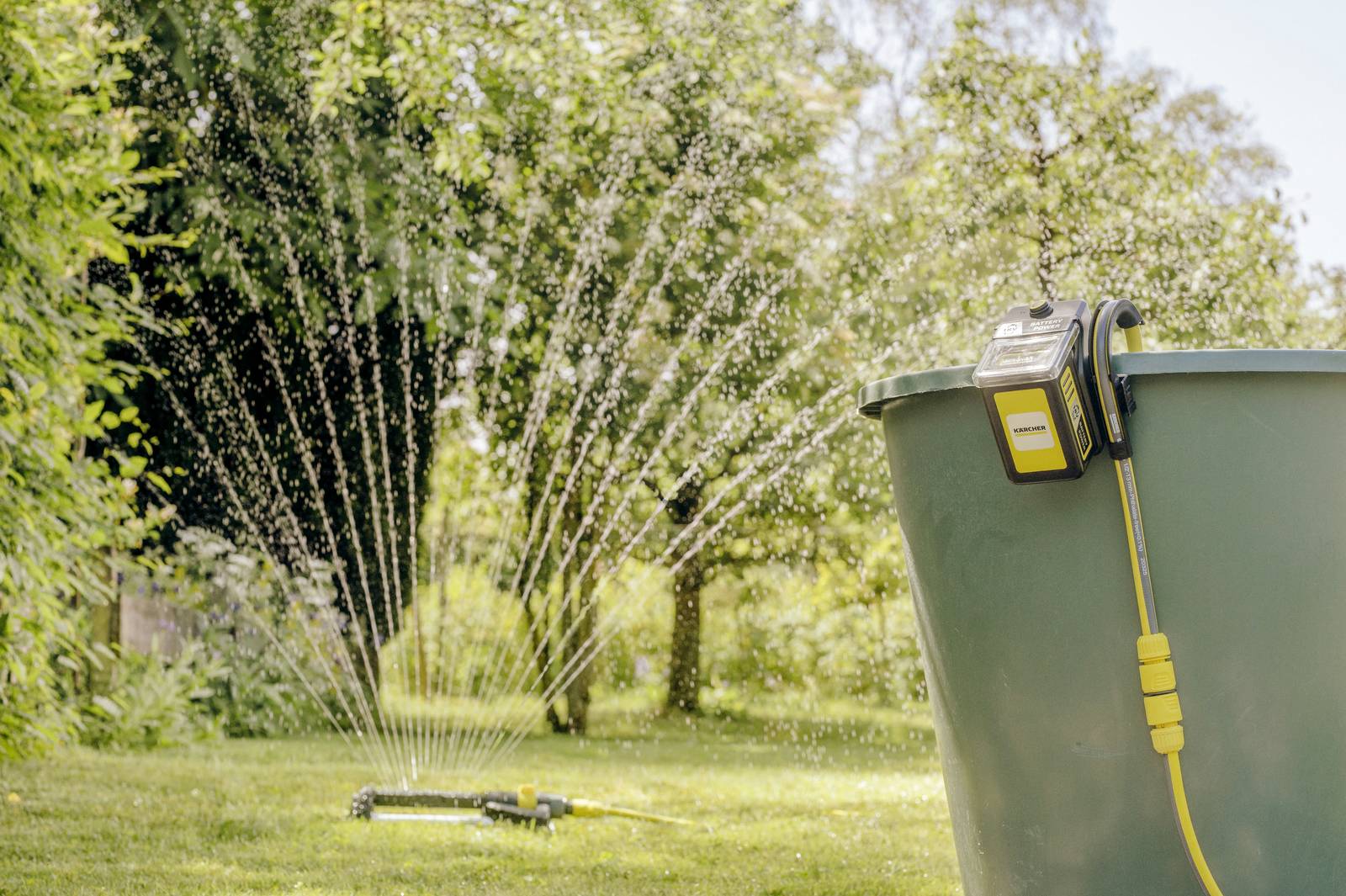 An irrigation system in the garden distributes water evenly across the lawn. In the foreground, there is a yellow container with a pump.