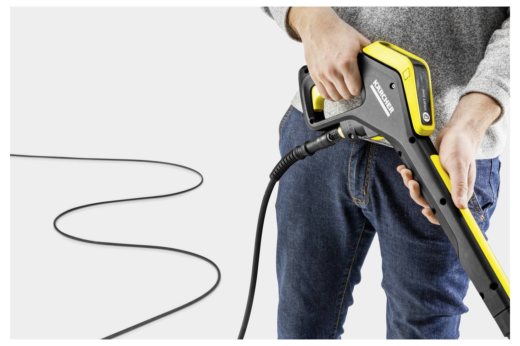 'A person holding a Kärcher pressure washer handle, with a long black cord extending across the floor, against a plain background.'