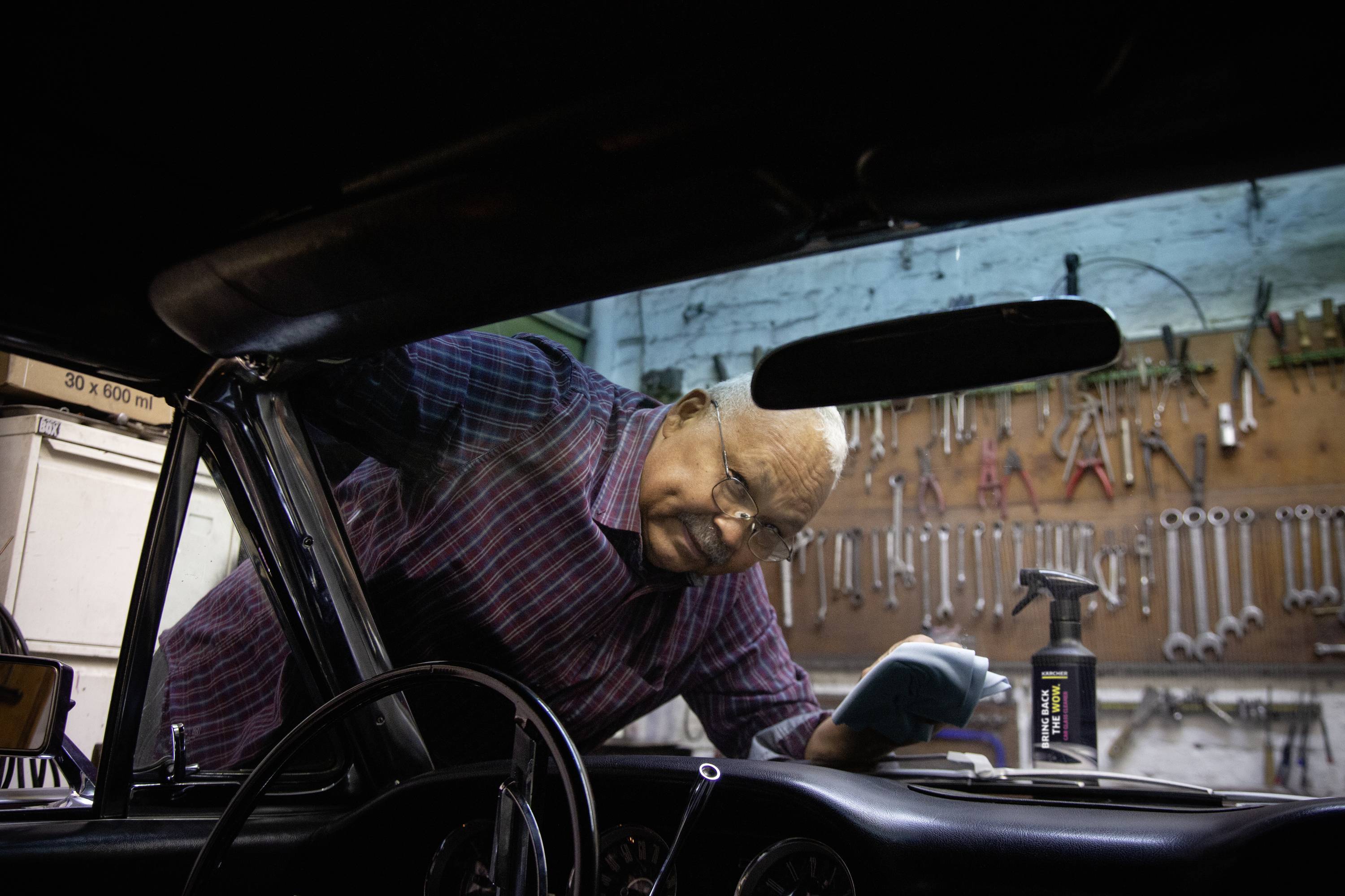 A man in a car is tinkering with the dashboard, with a workshop wall covered in tools in the background.