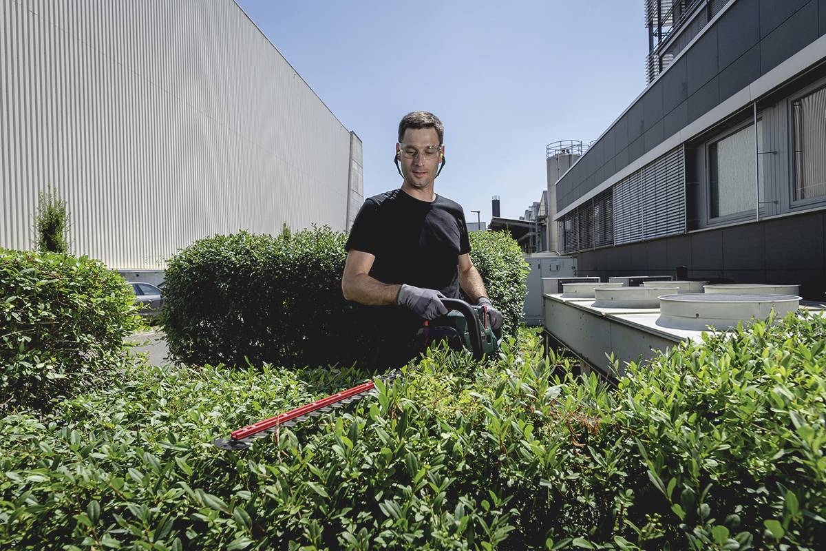 A man in a black T-shirt is trimming a hedge with hedge trimmers in front of a modern building.