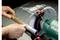 A person is holding a rusty pipe against a grinding machine. No sparks are flying, safety guard is in place. Metabo logo is visible.