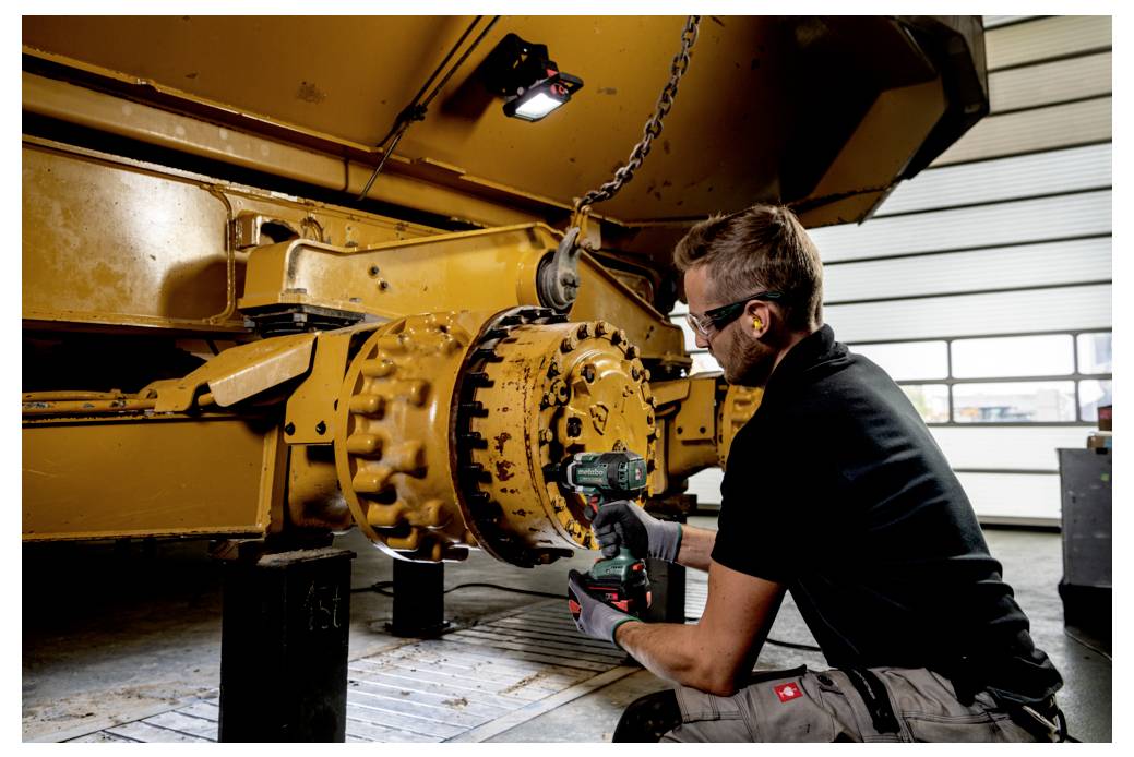 A worker uses a power tool to fix machinery in a workshop, wearing safety gear such as goggles and gloves. Machinery structure is visible.