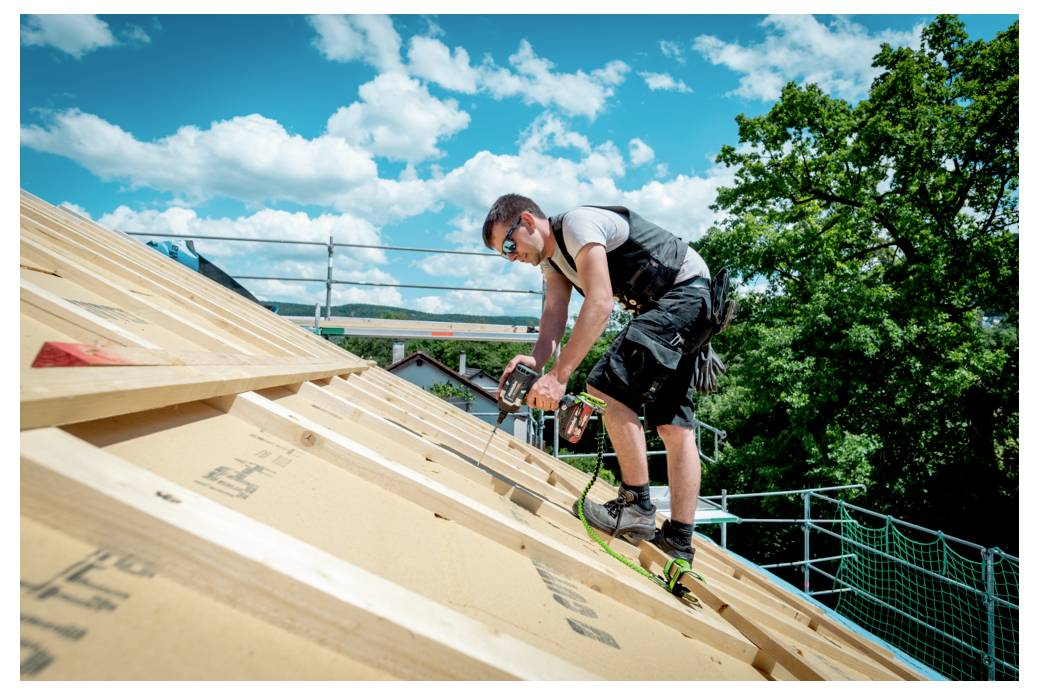 A construction worker in safety gear uses a nail gun on a wooden roof frame, set against a bright blue sky with clouds.