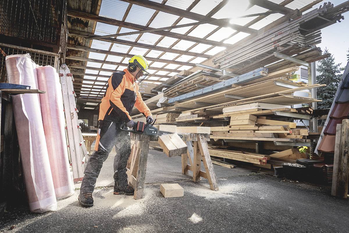 A person is sawing wood with an electric saw in a workshop. Stacked wooden boards and beams are visible in the background.