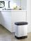 A metal bin stands on a light kitchen floor next to a white kitchen unit. Bananas and an orange are resting on the worktop.