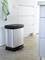Metal bin with a black lid and pedal stands in a modern kitchen on a stone floor. Towels are hanging to the right.
