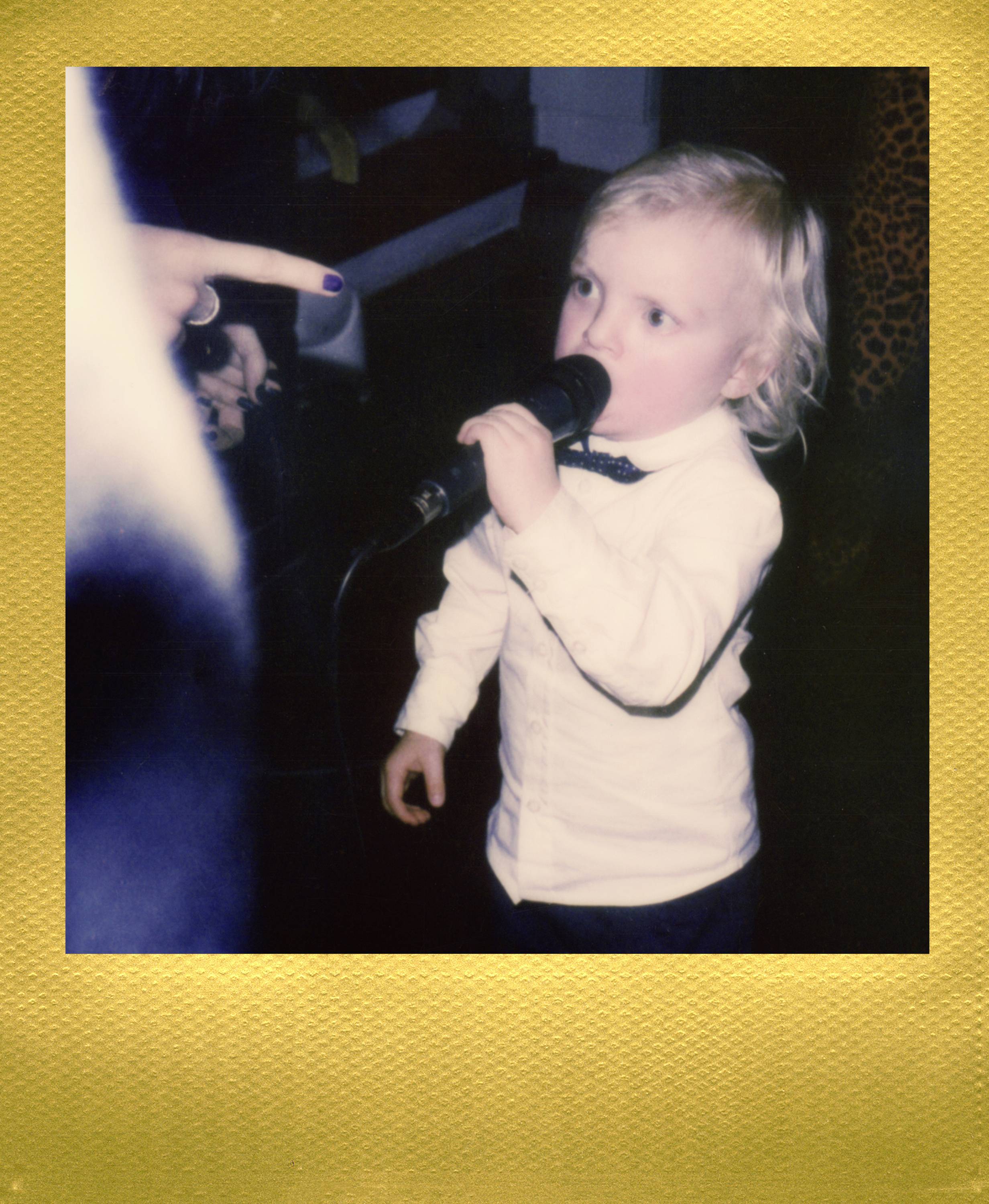 A small child in a white shirt is holding a microphone and speaking into it, while a hand points at the child.