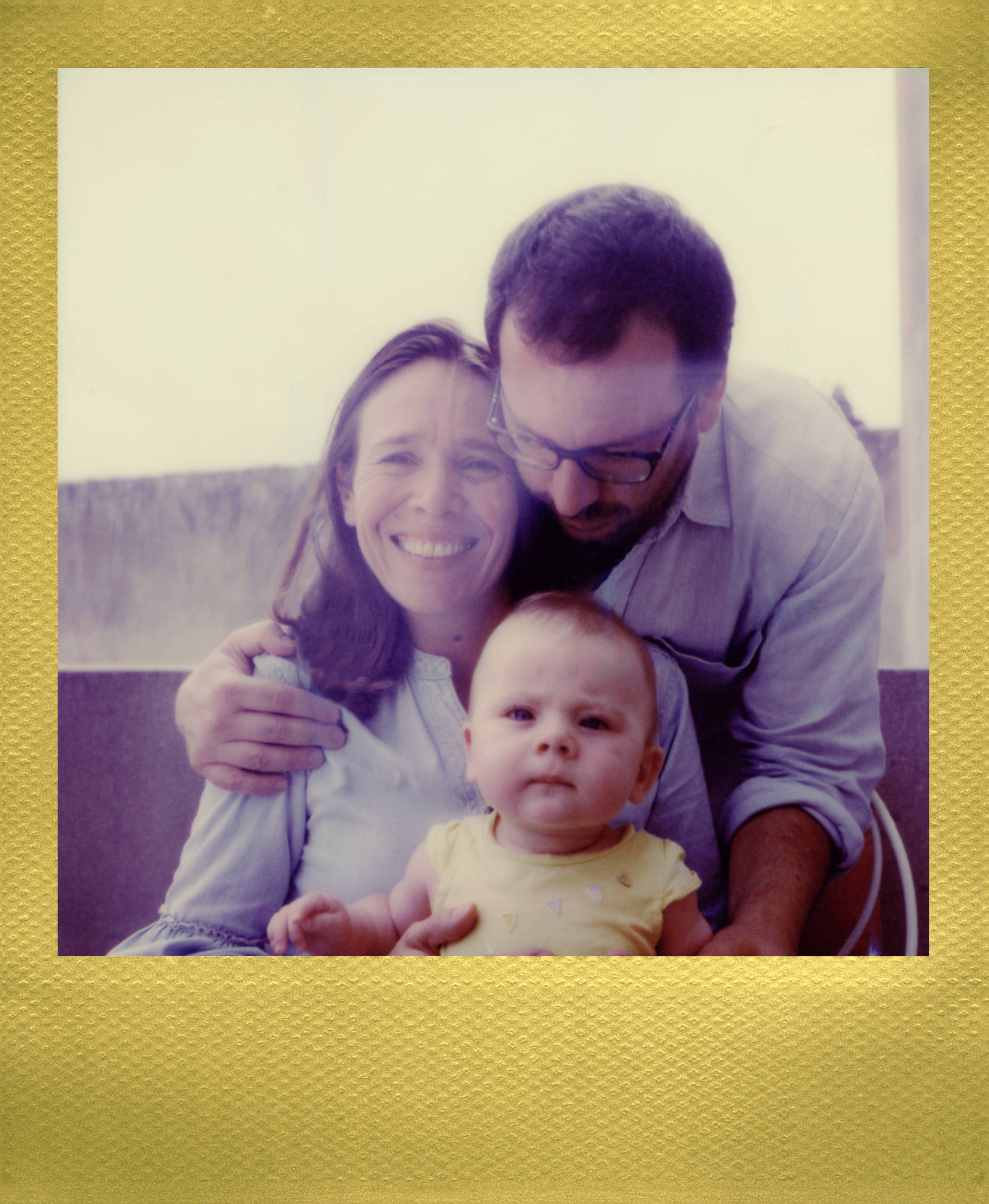A woman and a man are sitting with a baby on their lap. The woman is smiling at the camera, while the man looks down at the baby.