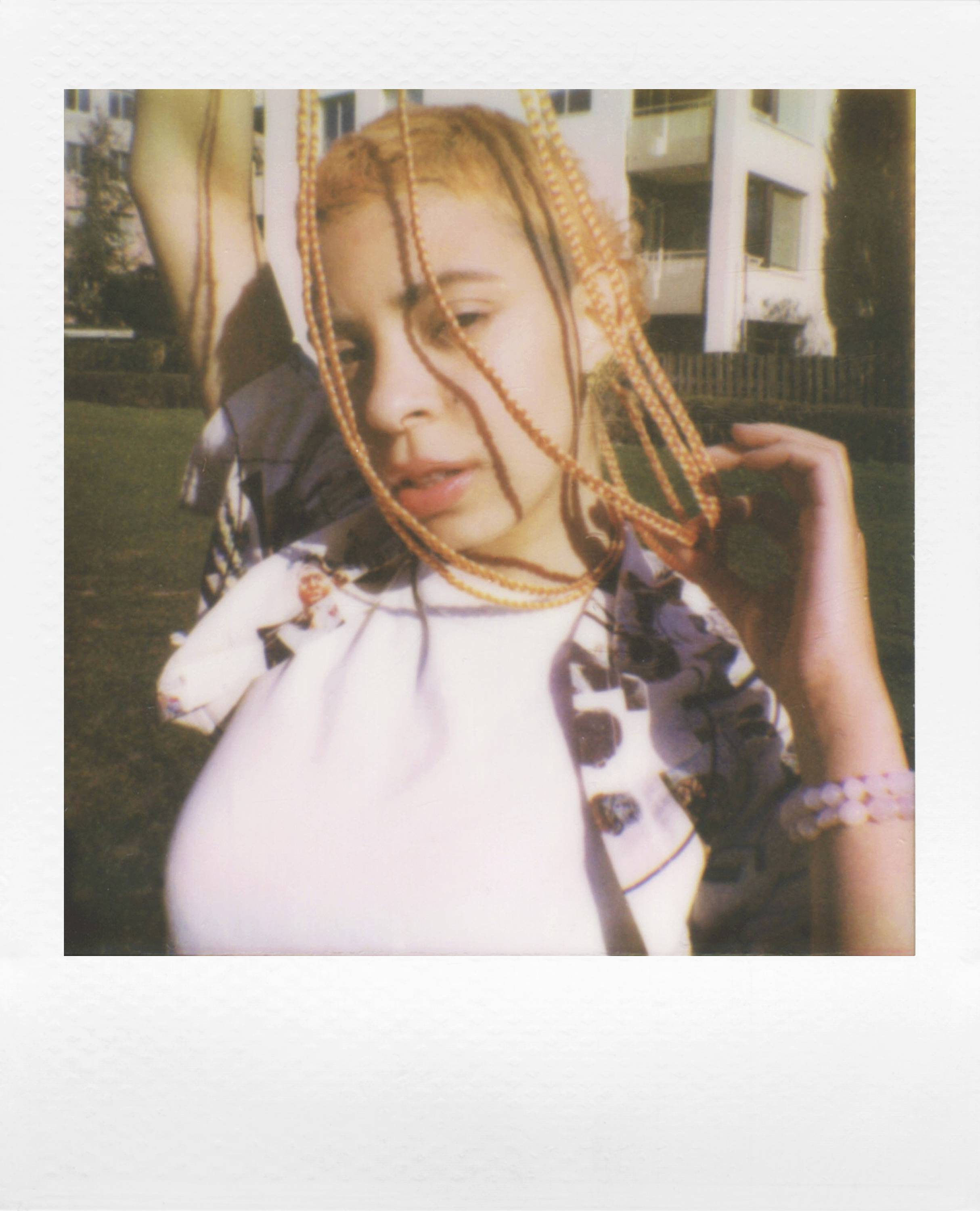 A person with colourful hair is posing outdoors in front of a building. They have one hand on their head and are wearing a white top.