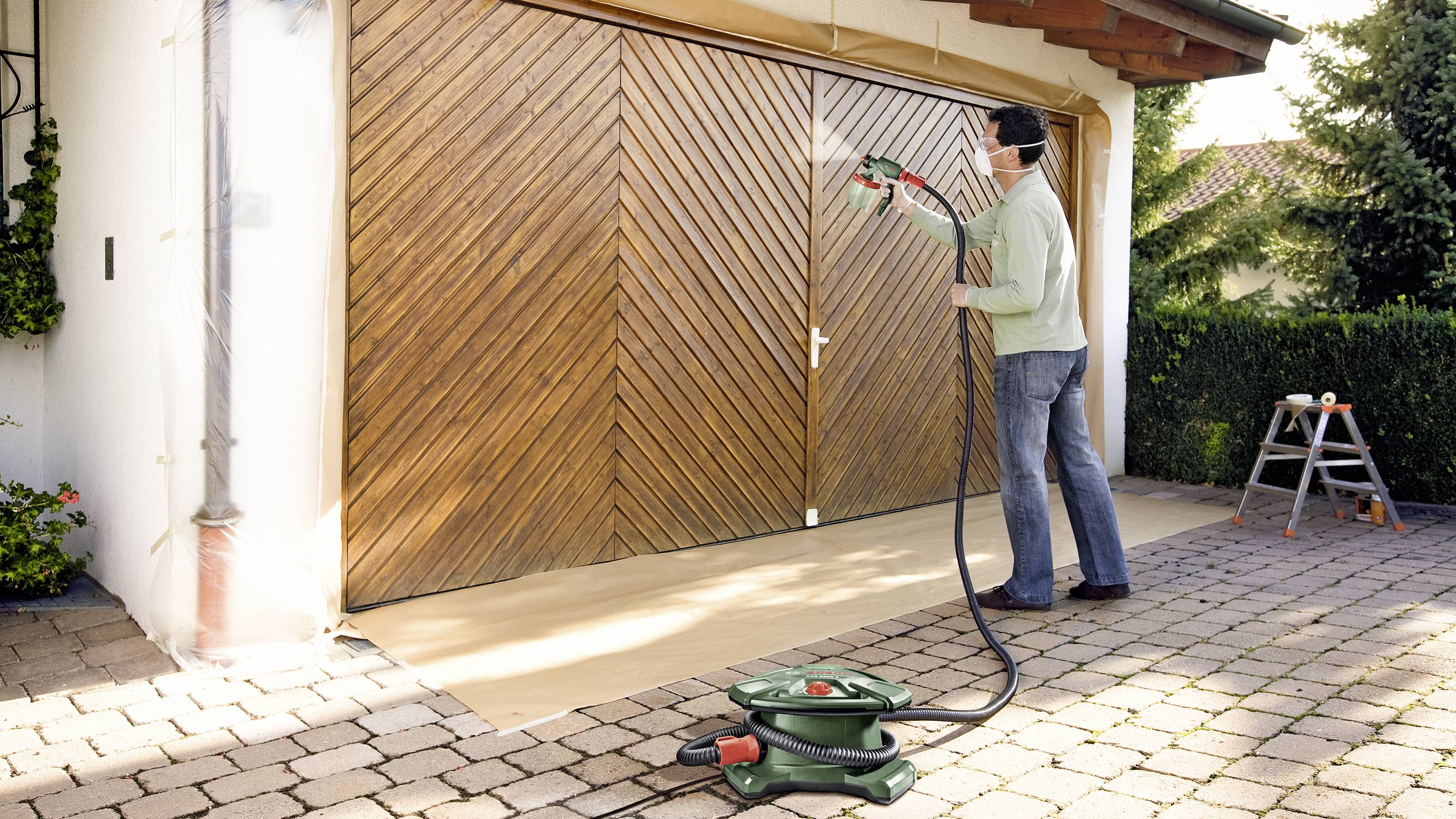 A person is spraying wood paint onto a large garage door using a spray painting system. A house and garden are visible in the background.