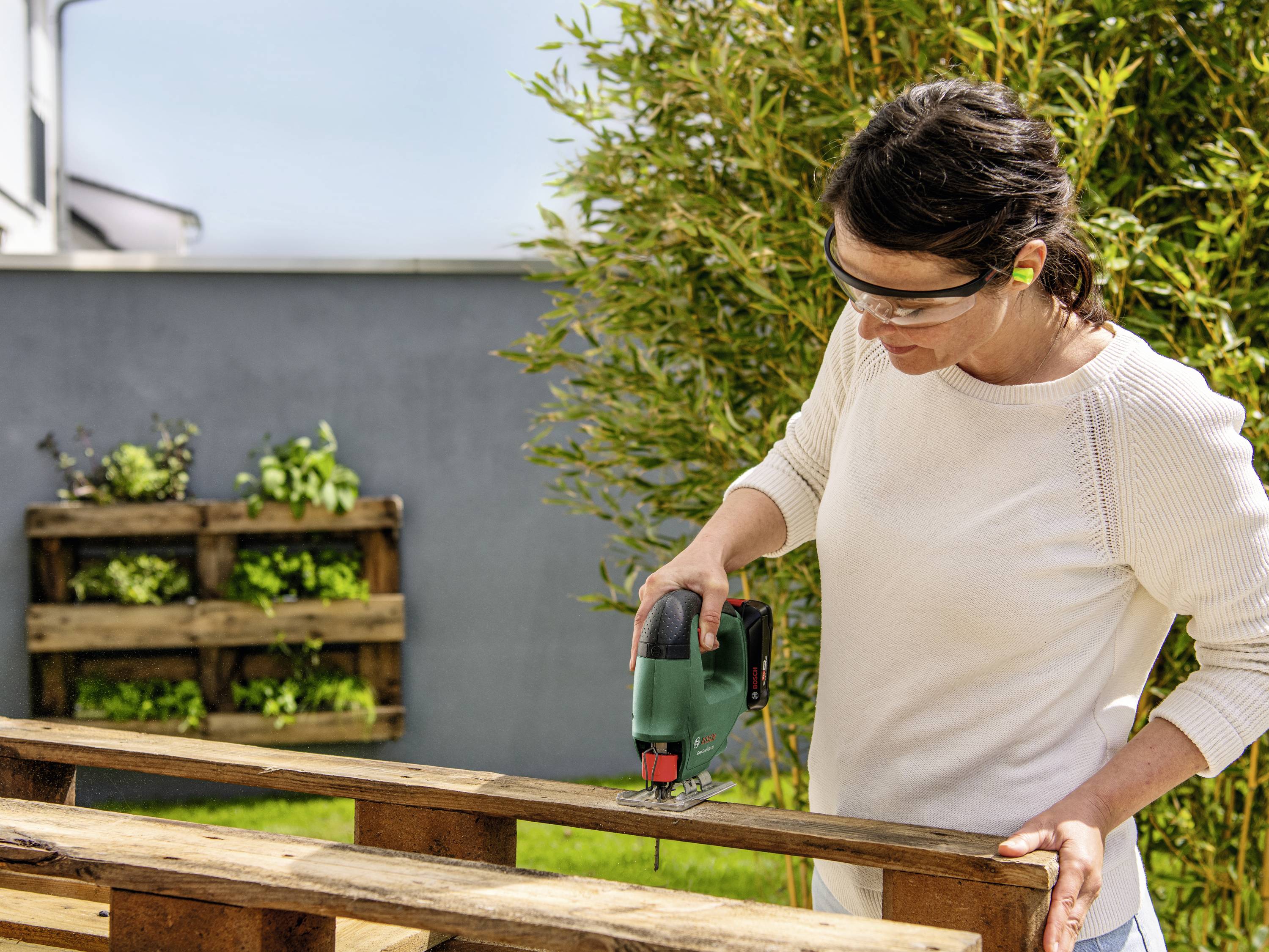 A person is sawing an outdoor wooden pallet using an electric saw. They are wearing safety glasses and ear protection. Plants can be seen in the background.