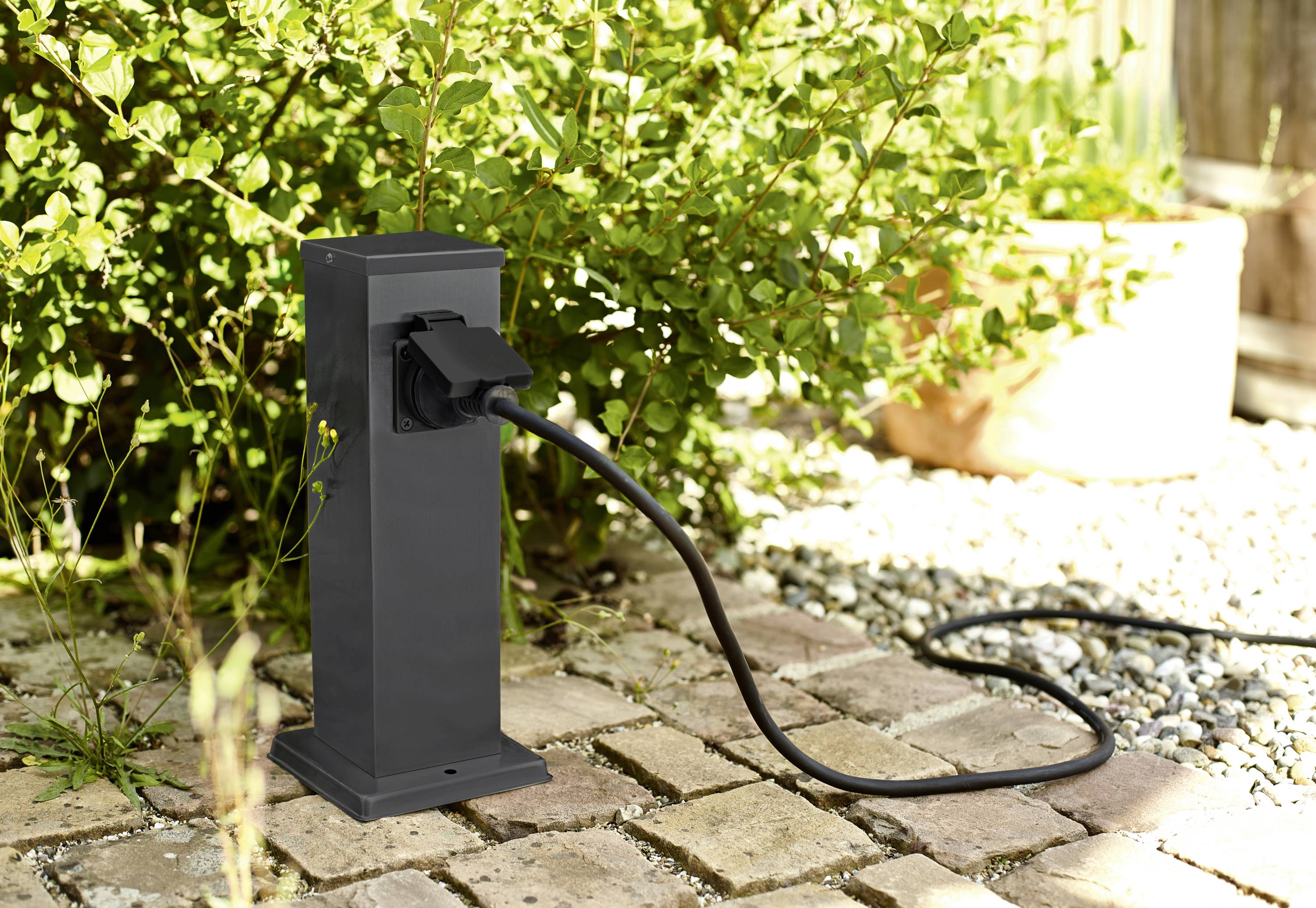 A black outdoor electrical column with an attached cable stands on paving stones in front of a planted background.