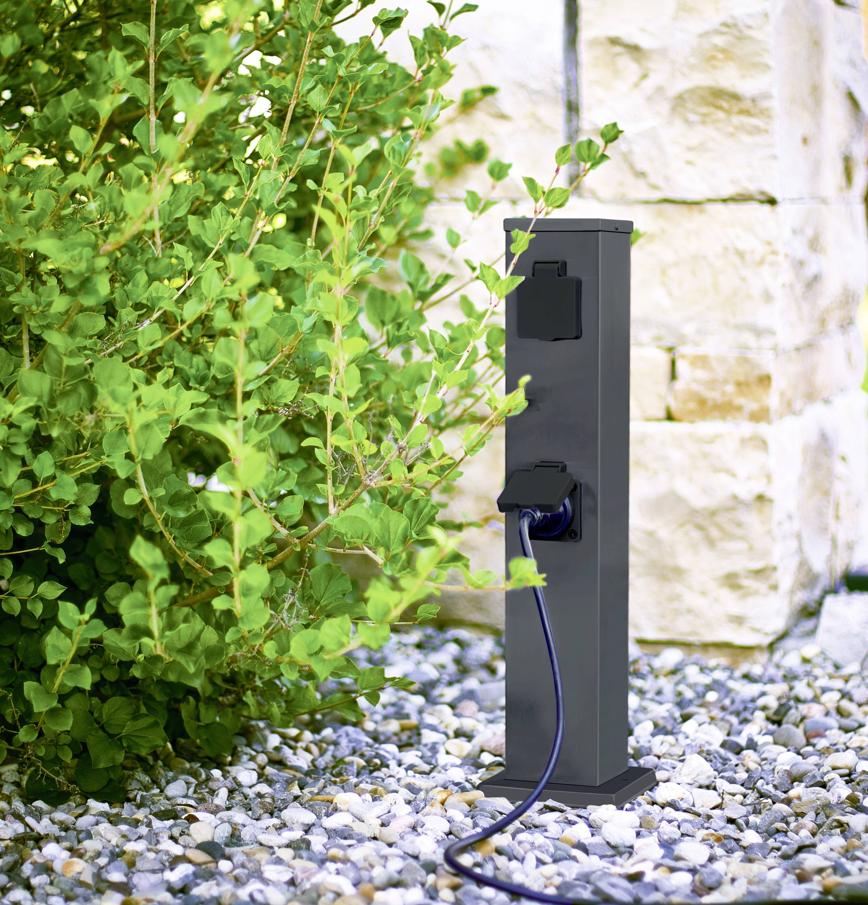 A black outdoor socket stands in a gravel bed next to a green bush. A cable is connected to the socket.