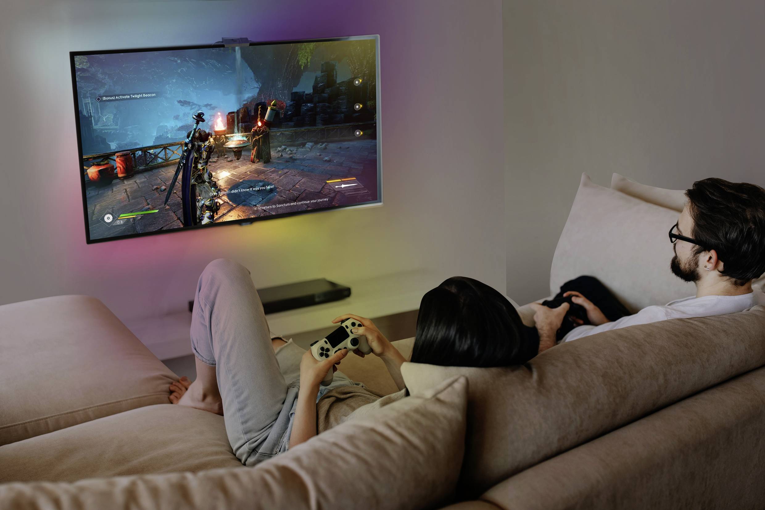 A woman and a man are sitting on a sofa, playing a video game on a large television mounted on the wall. They are using game controllers.