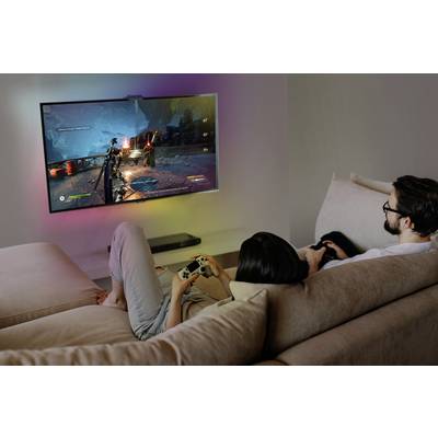 A woman and a man are sitting on a sofa, playing a video game on a large television mounted on the wall. They are using game controllers.