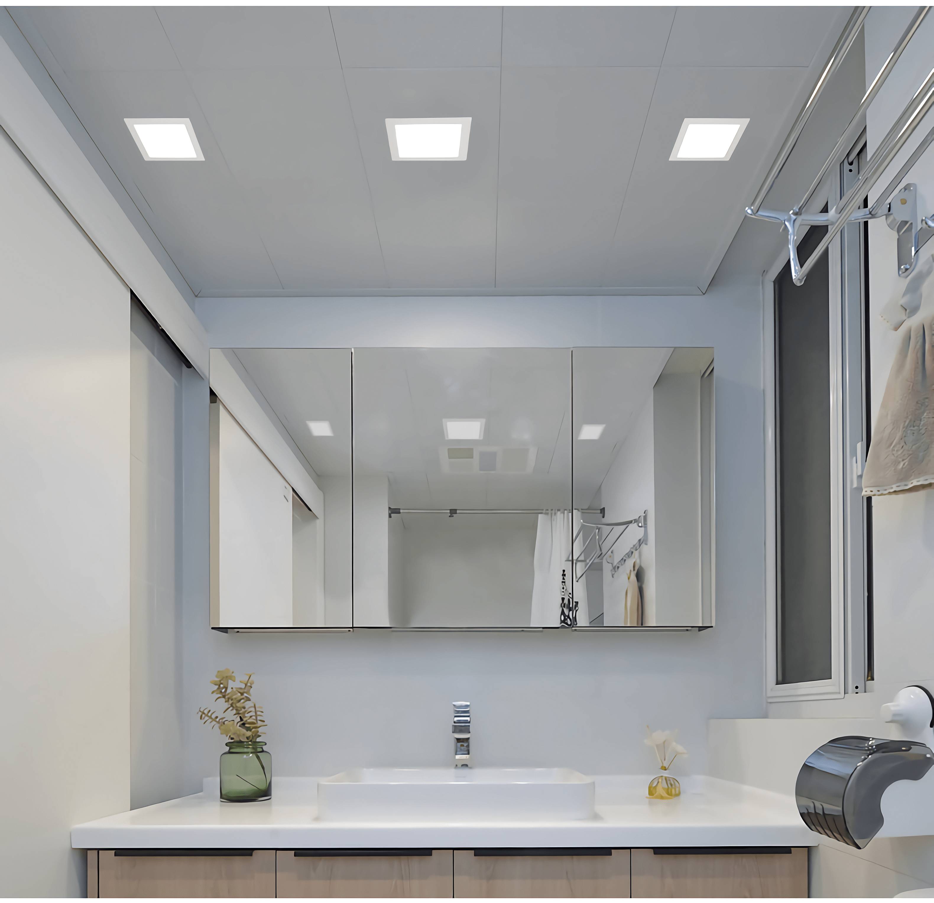 Modern bathroom with a large mirror, washbasin, plant in a glass vessel and towel on a hook. Ceiling lights illuminate the room.