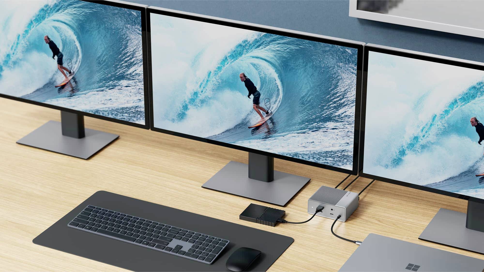 Three computer monitors on a desk display surfers riding a large wave. A keyboard and mouse are positioned in front of them.