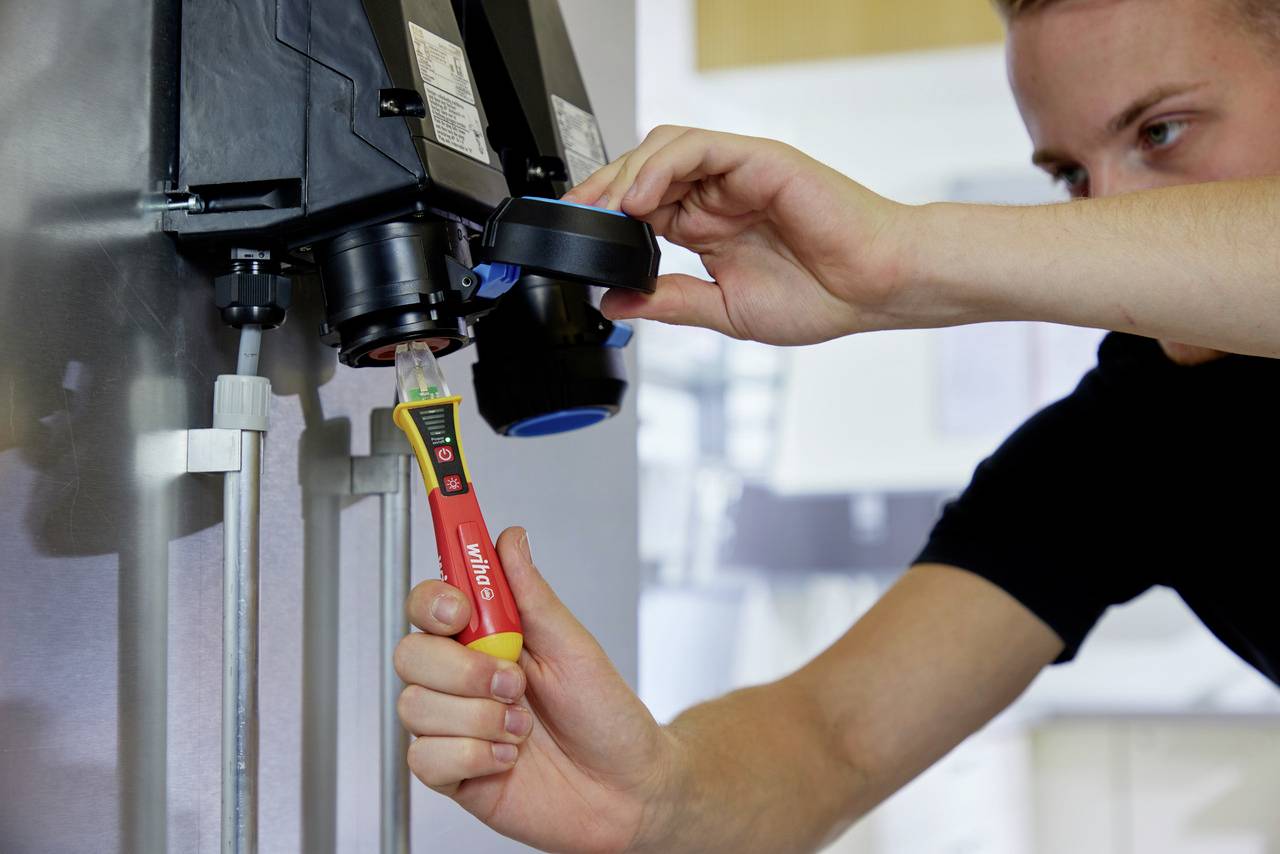 A man is measuring the voltage at an electrical connection using a voltage tester. He is wearing a black shirt.