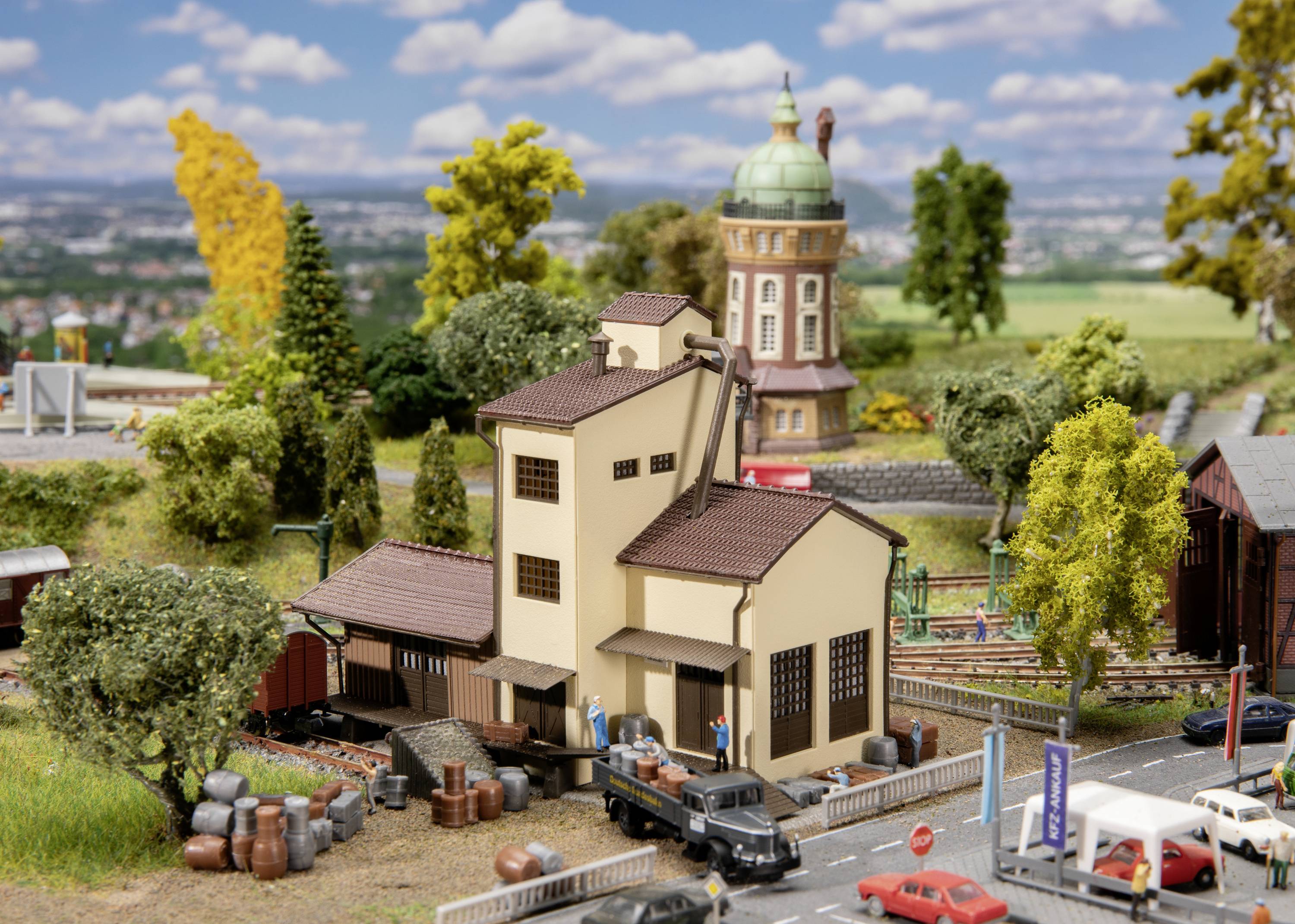 Miniature landscape featuring a main building, surrounded by trees and a water tower in the background. A lorry and people are in the foreground.