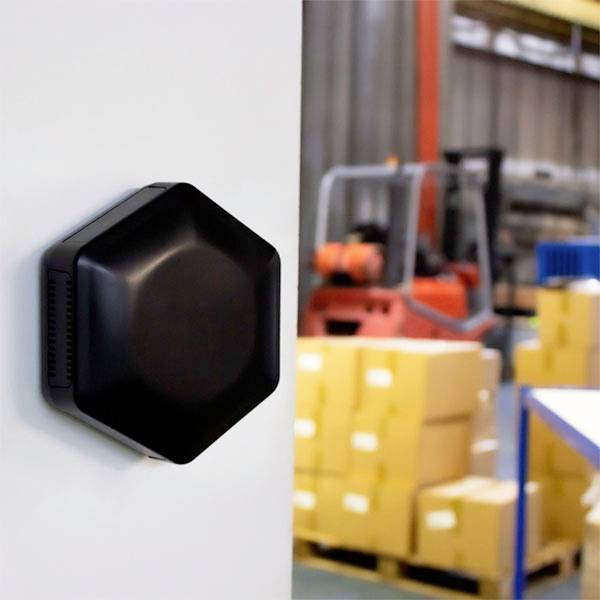 A black, hexagonal sensor is mounted on a white wall. Cardboard boxes and a blurred machine can be seen in the background.